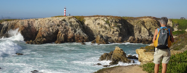 Individuele wandelreis Rota Vicentina, Portugal Vrouw met rugzak staat op kliffen aan zee met vuurtoren op de achtergrond, in Portugal.